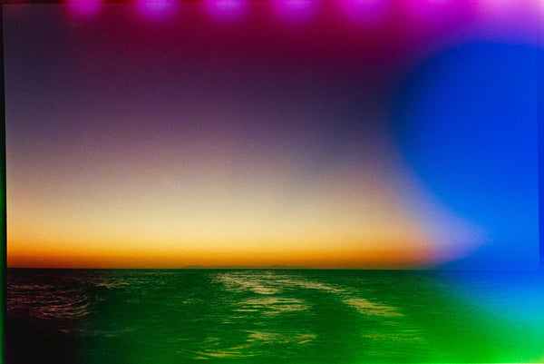 A colorful landscape photograph of an ocean horizon at sunset or sunrise, with a purple-to-orange gradient sky and green-blue water. The image has a strong blue vignette on the right and a green tint along the bottom edge.