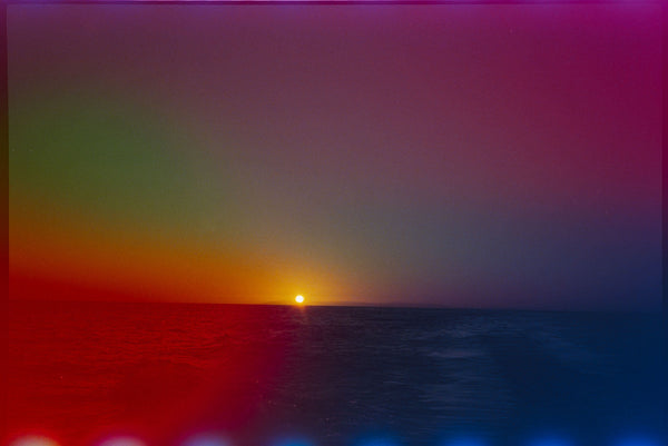 A colorful sunset seascape with the sun low over the horizon, showing a gradient sky from green and orange on the left to purple and deep blue on the right, with a red reflection on the water.