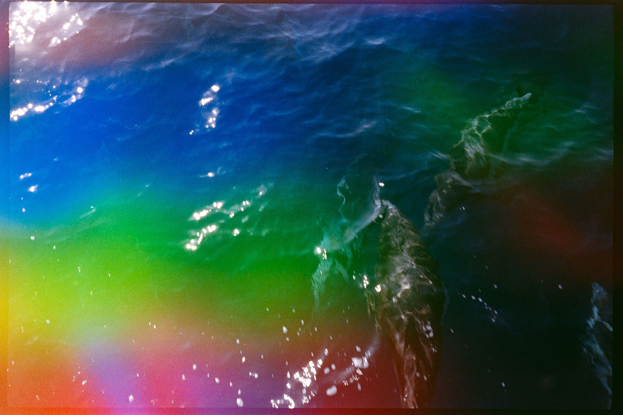 A colorful abstract seascape photograph showing rippling ocean water with strong blue, green, yellow, pink, and orange tones, plus bright sunlight reflections.