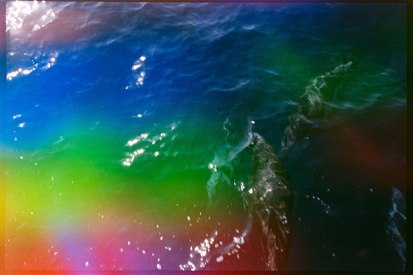 A colorful abstract seascape photograph showing rippling ocean water with strong blue, green, yellow, pink, and orange tones, plus bright sunlight reflections.
